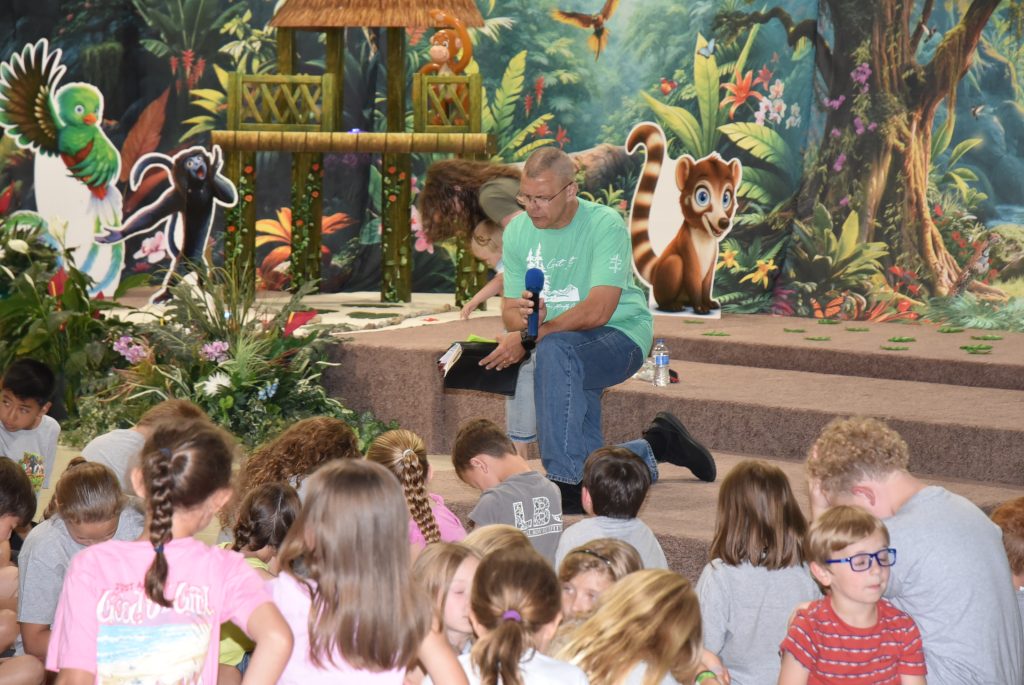 A man sits on stage with a microphone, surrounded by young children, in front of a colorful jungle-themed backdrop featuring animals and plants.