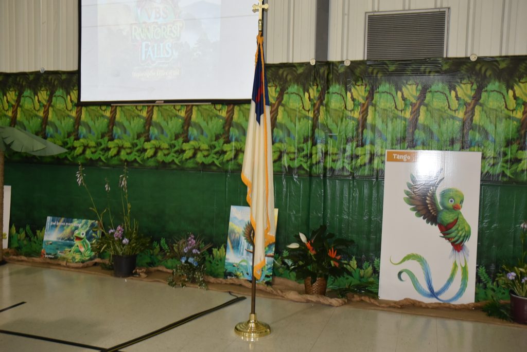 A flag on a stand in front of a jungle-themed backdrop with plants, a screen, and a sign featuring an eagle emblem.