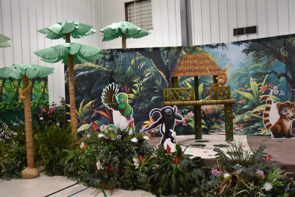 A jungle-themed indoor display with artificial palm trees, plants, a thatched hut, and cartoon animal cutouts in front of a rainforest backdrop.