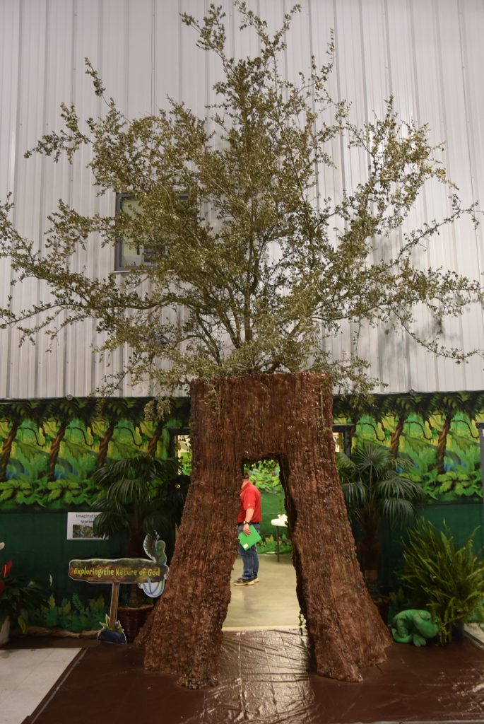 A person in a red shirt walks through a large artificial tree structure indoors, with plants and a mural in the background.