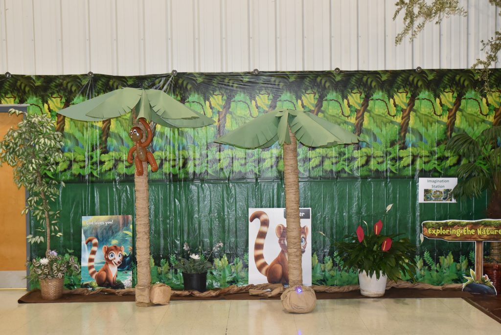 Jungle-themed party decoration with two artificial palm trees, cartoon lemur images, green backdrop, and potted plants.