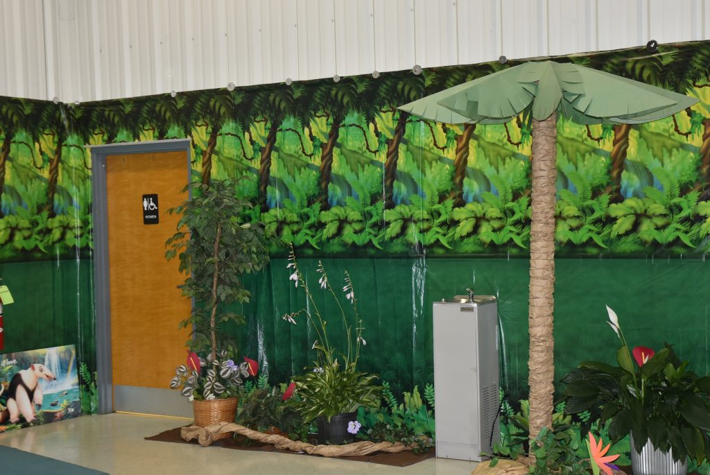 Jungle-themed decor with artificial trees and plants next to a water fountain and restroom door, set against a jungle mural wall.