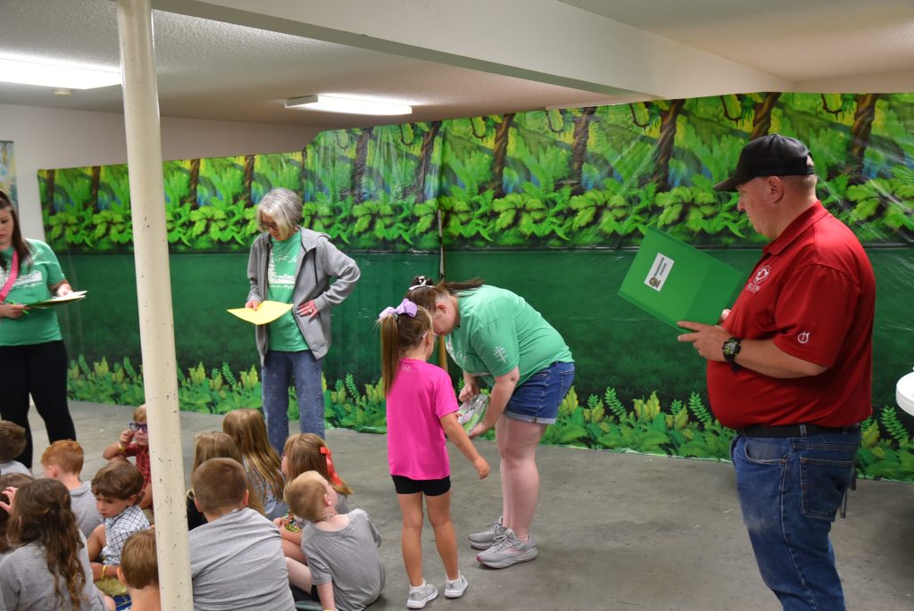 Adults and children gather in a room with jungle-themed wall decorations; an adult hands something to a child while others watch.