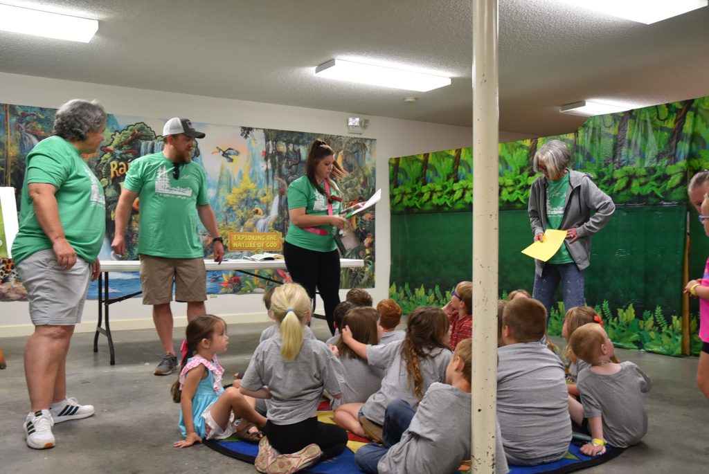 Three adults stand in front of a seated group of children indoors, holding papers, with colorful murals on the walls in the background.