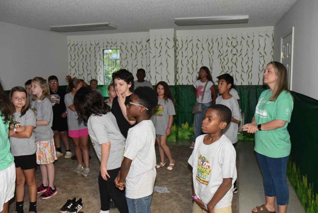 A group of children and adults stand together inside a room with painted walls, some engaging in conversation and activities.