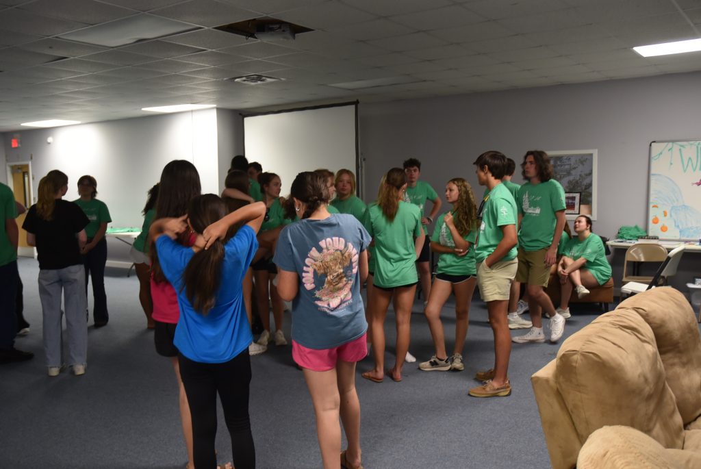 A group of young people in casual clothes gather and talk in a large room with gray walls and a projector screen in the background.