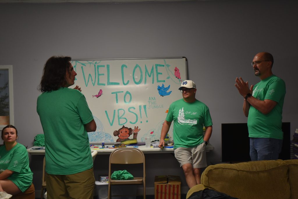 Three people in green shirts stand near a "Welcome to VBS!!" sign in a decorated room, with a child sitting at a table beneath the sign.