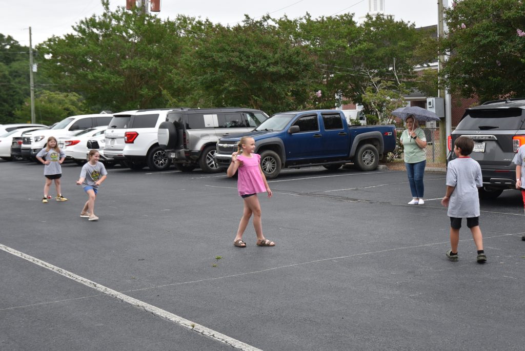 Children and adults stand and walk in a parking lot with several parked vehicles in the background.