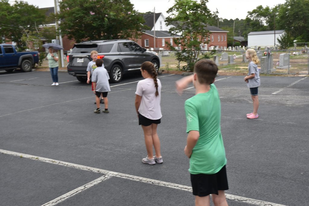 Several children stand and play outside on a paved area, near parked cars and trees, on a cloudy day.