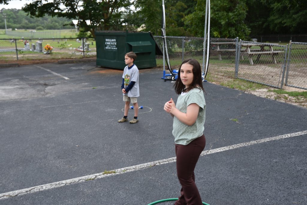 Two children playing outside on a paved area near a green dumpster, with trees and a metal fence in the background.