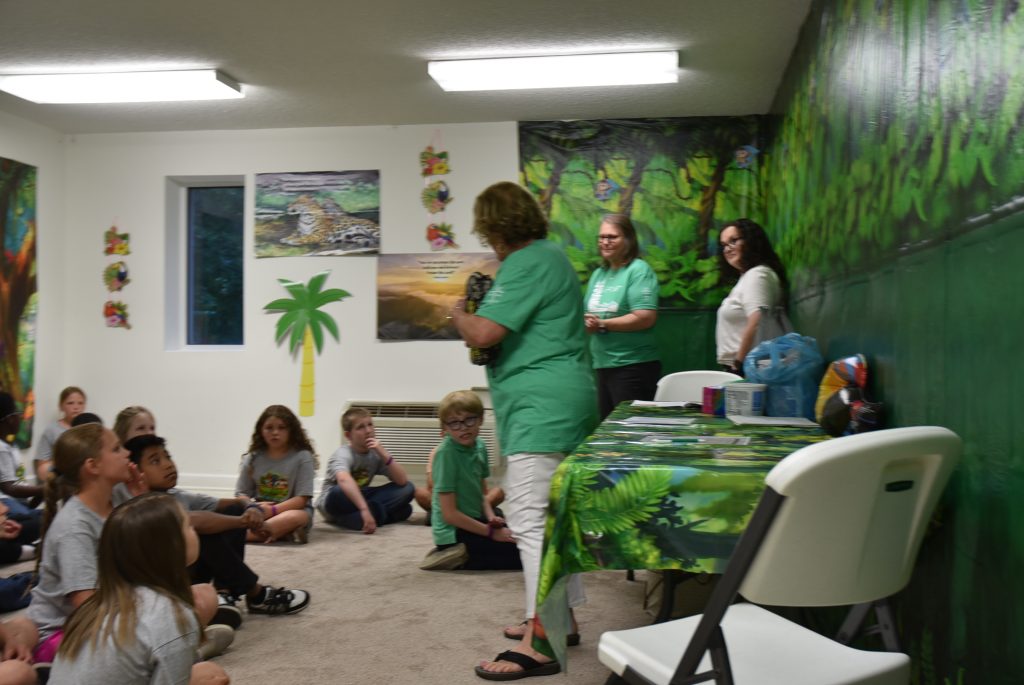 A group of children sits on the floor while three adults stand and talk in a brightly decorated classroom with a jungle theme.