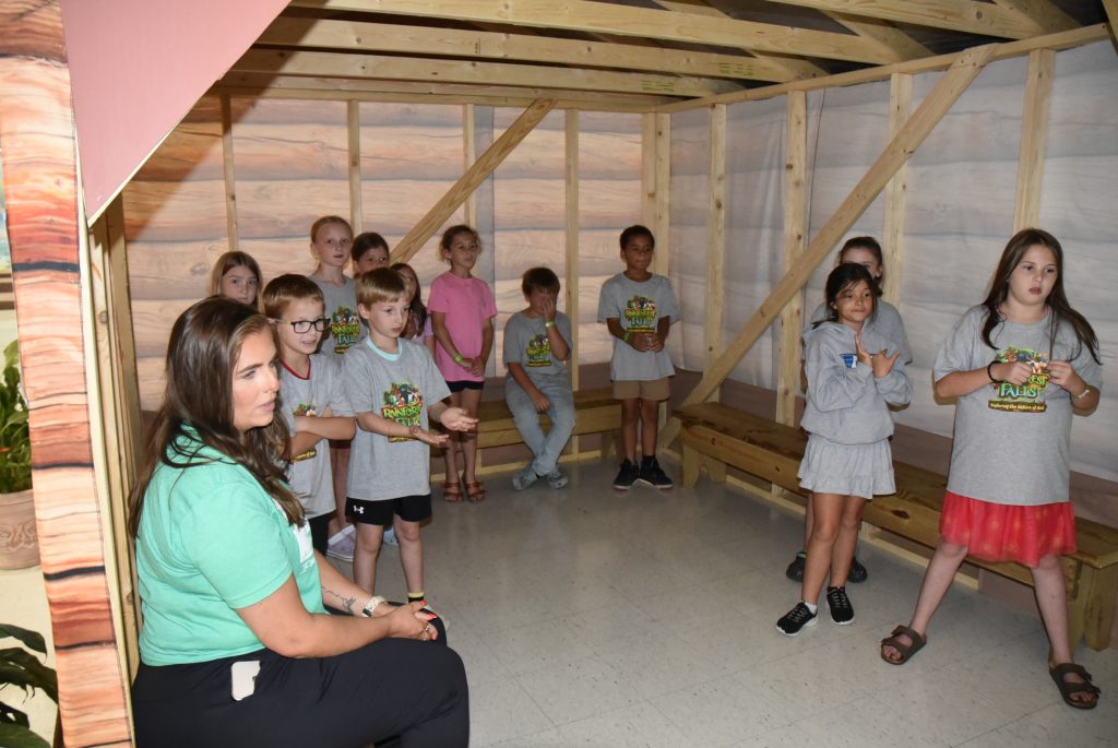 A group of children and an adult sit and stand inside a wooden structure, possibly a classroom or activity area, listening and interacting together.