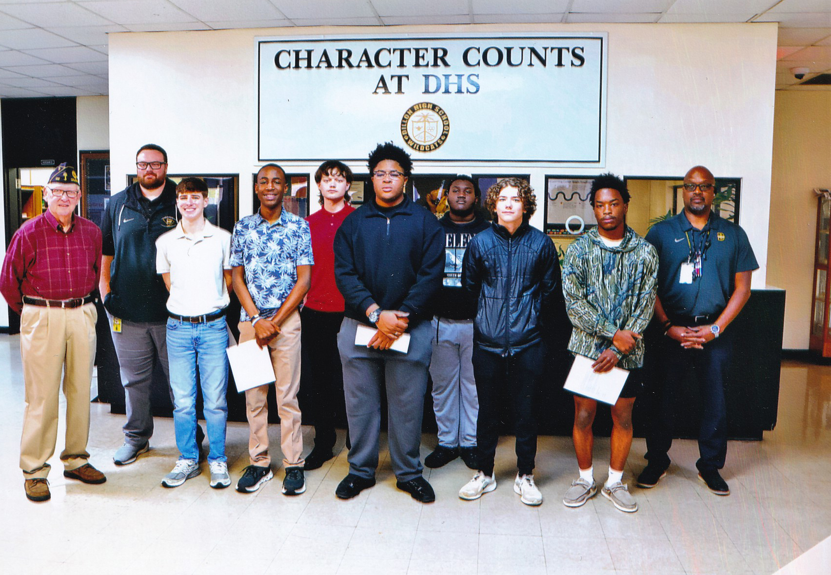 A group of students and staff stand in front of a wall sign reading "Character Counts at DHS" inside a school hallway.