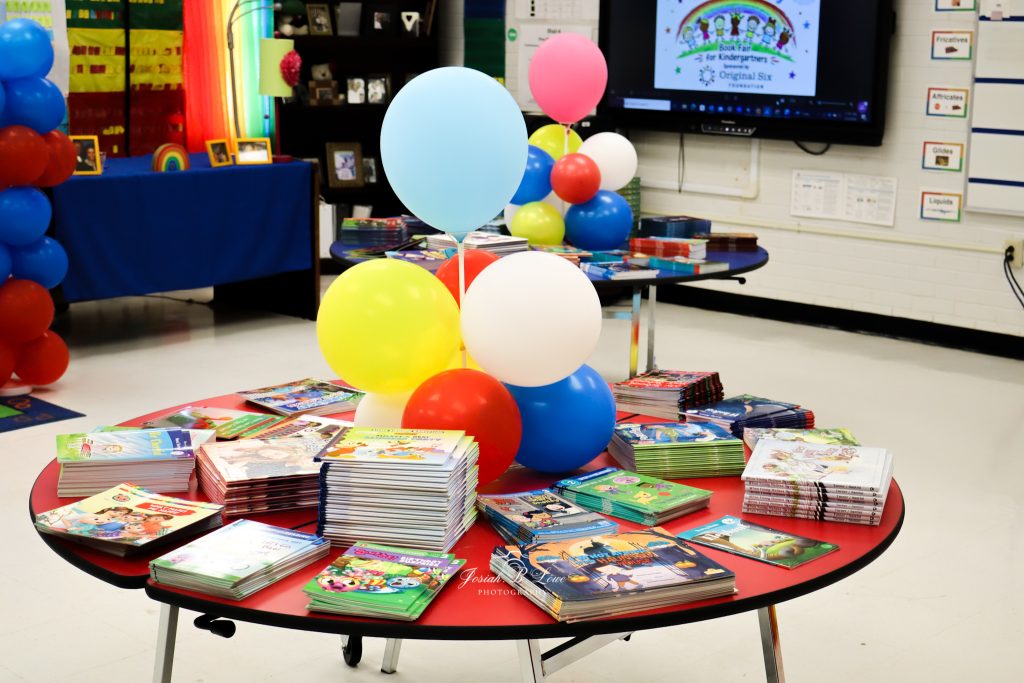 Stacks of colorful children's books on round tables with balloons, set up in a classroom for a book fair.