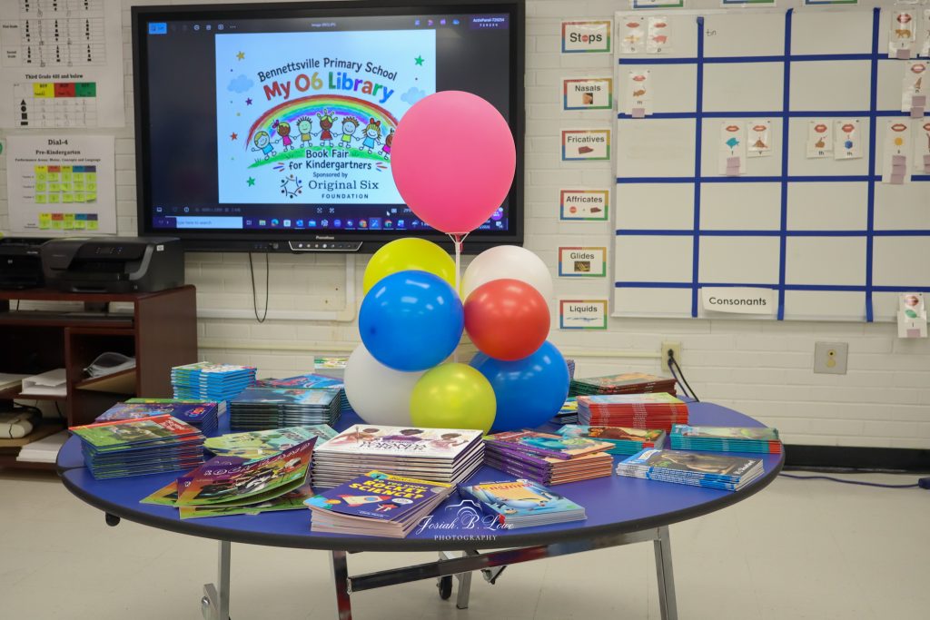 Colorful books and balloons arranged on a round table in a classroom, with a screen displaying "Book Fair for Kindergartners" in the background.