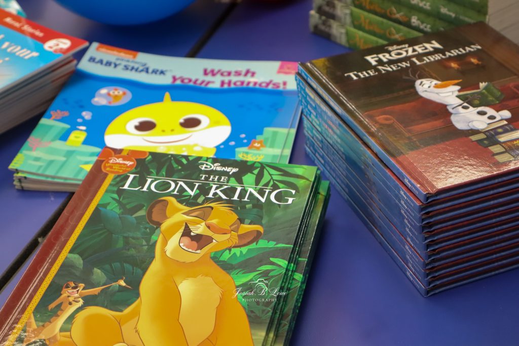 Stacks of children's books, including "The Lion King," "Frozen: The New Librarian," and "Baby Shark: Wash Your Hands!" displayed on a blue table.