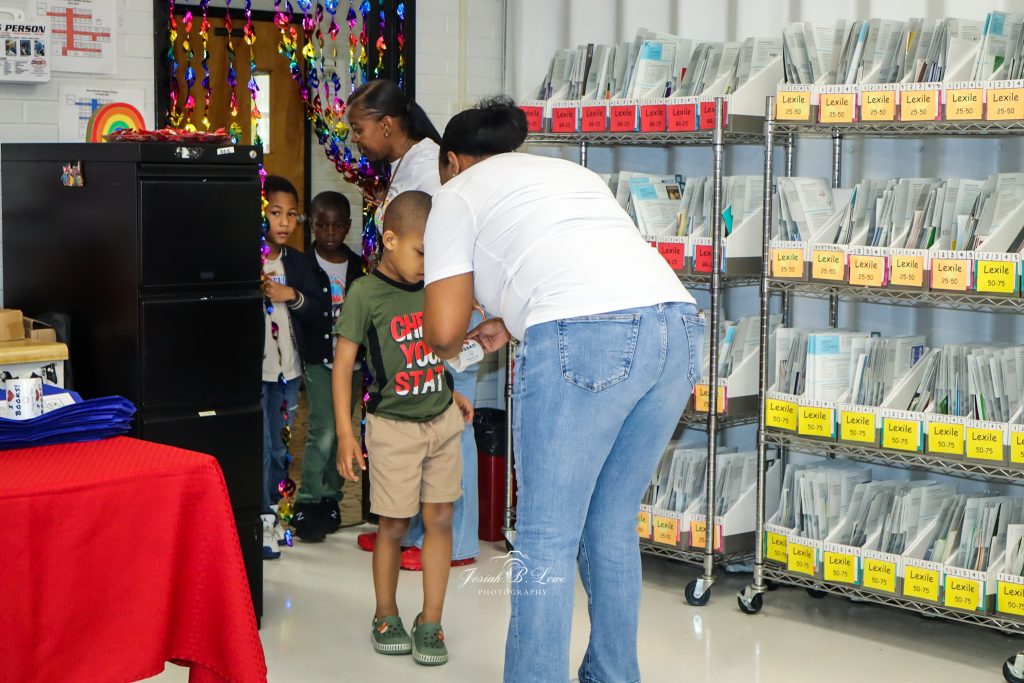 A teacher greets young children entering a classroom with bookshelves labeled by reading levels and colorful decorations on the doorway.
