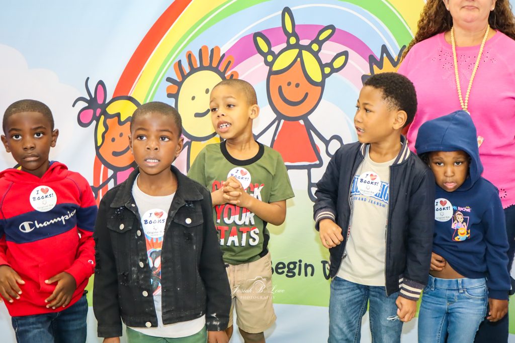 Five young boys stand in front of a colorful mural with rainbows and cartoon children, wearing “I love books” stickers, with a partially visible adult behind them.