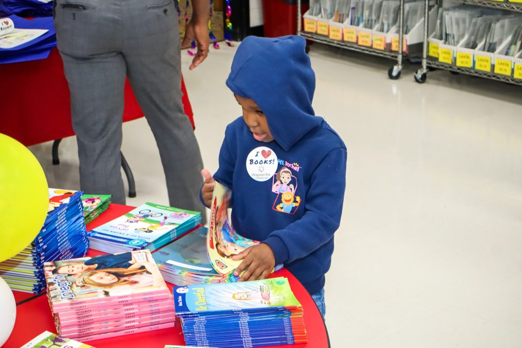 A young child in a blue hoodie looks at a book while standing by a table stacked with colorful children's books in a classroom or library setting.