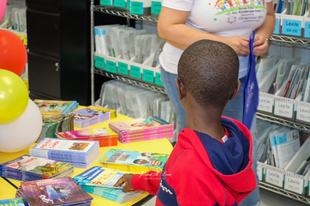 A child in a red hoodie looks at a table filled with colorful books, while an adult stands nearby in a classroom or library setting.