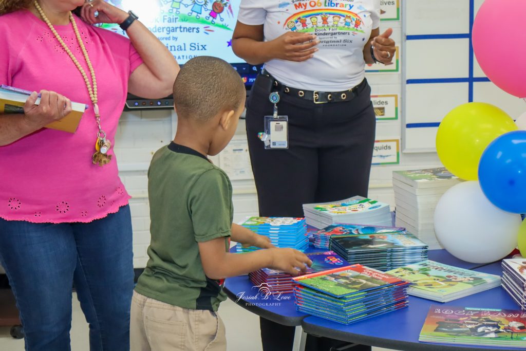 A young boy at a book fair selects books from a table, with adults and colorful balloons nearby.