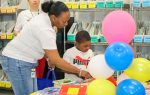Woman and child look at books on a table with colorful balloons in a library or classroom, shelves labeled by reading levels in the background.