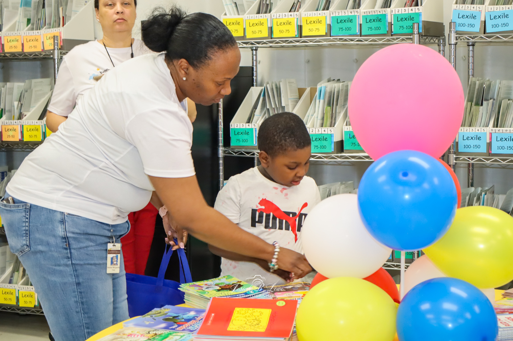 Woman and child look at books on a table with colorful balloons in a library or classroom, shelves labeled by reading levels in the background.