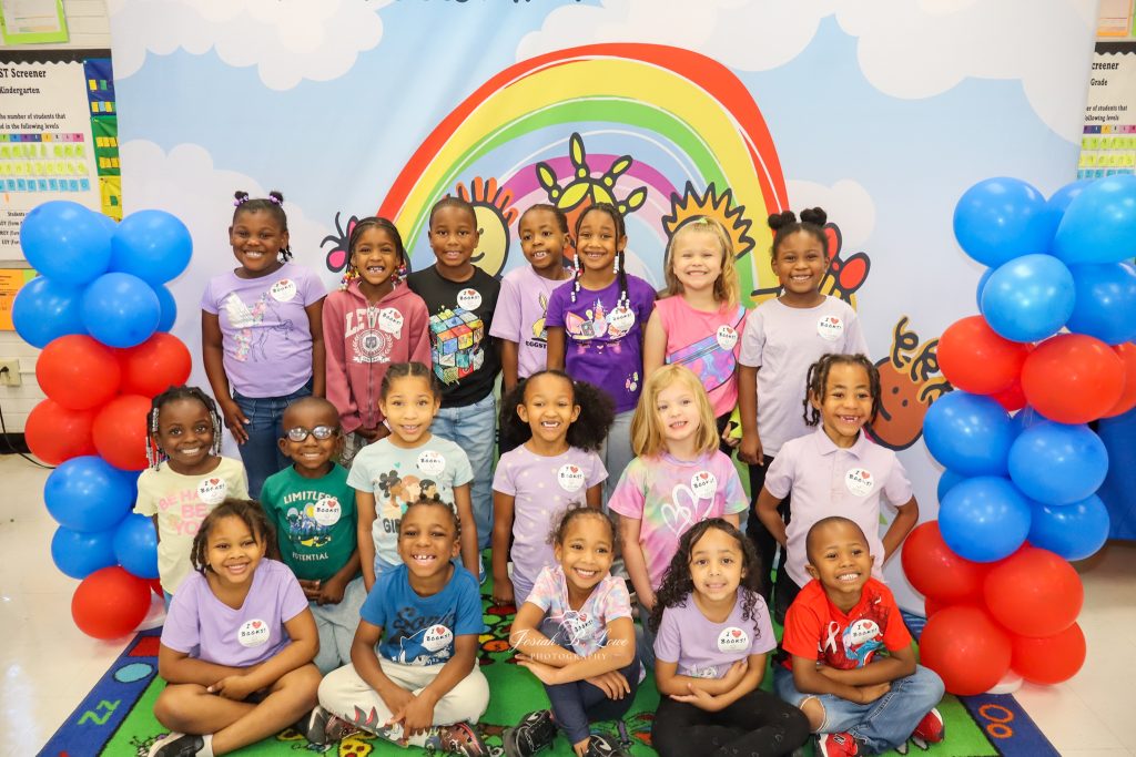 A group of diverse young children pose smiling in front of a colorful rainbow backdrop, with red and blue balloon columns, in a classroom setting.