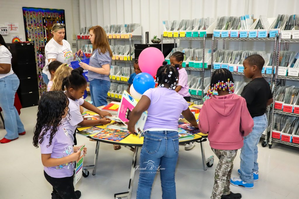 Children and adults gather around a table of books in a library, looking at reading materials, with bookshelves and colorful balloons in the background.