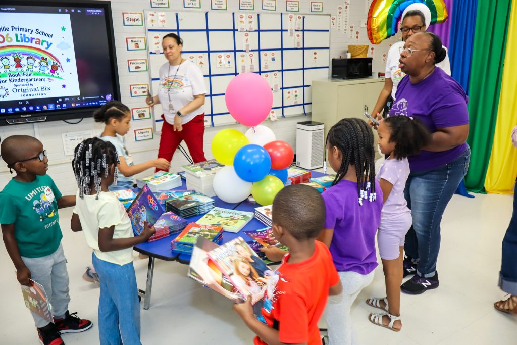 Children and adults gather around a table with books and balloons at a school book fair in a colorful classroom setting.