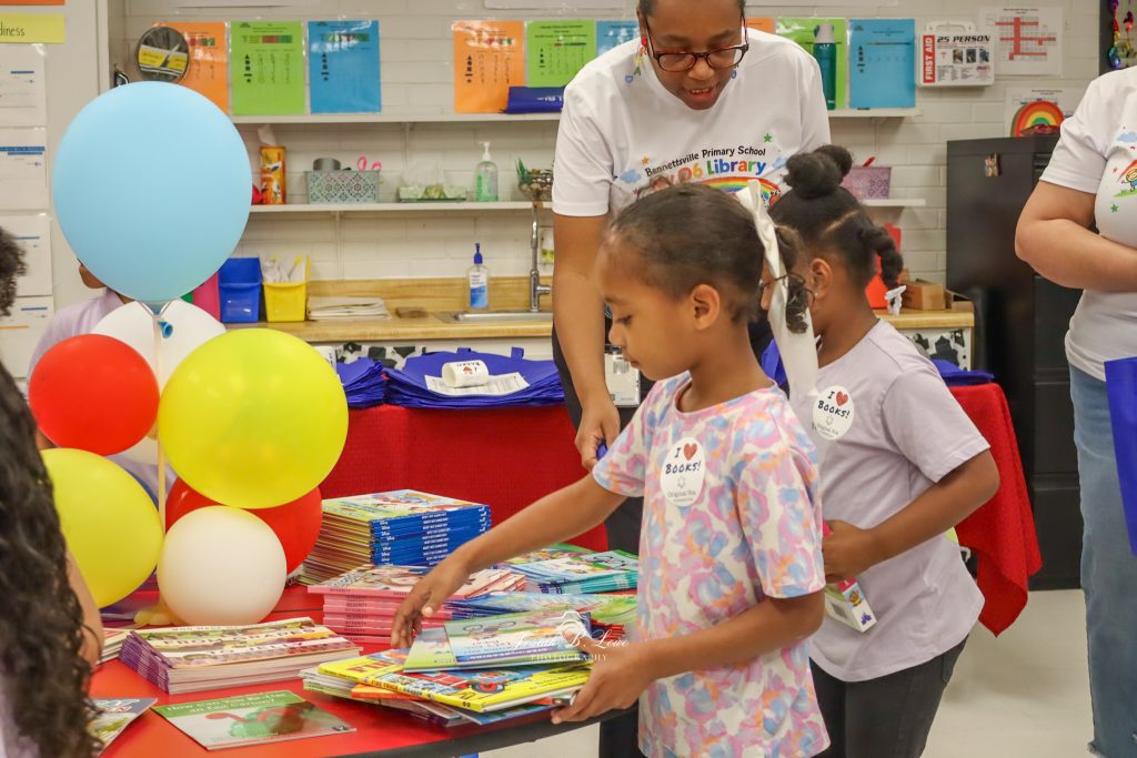 Young children select books from a table at a school library event, with colorful balloons and adults assisting in the background.