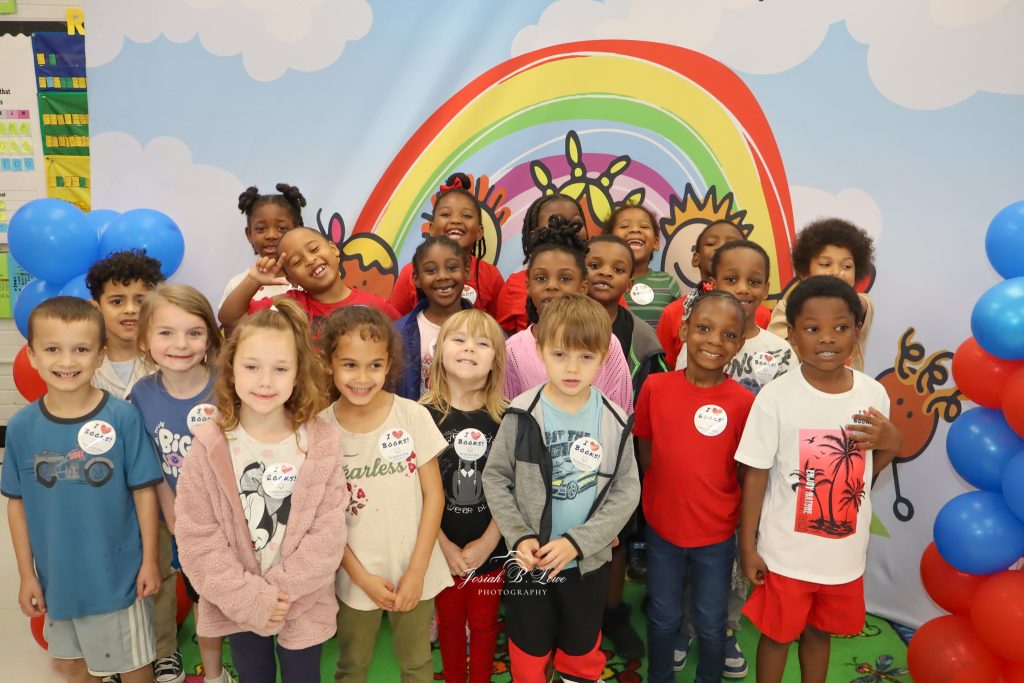 A group of smiling children stand together in front of a colorful rainbow backdrop, surrounded by blue and red balloons.