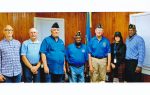 Seven people stand side by side in a room with wooden walls, some wearing military hats and blue shirts, posing for a group photo.