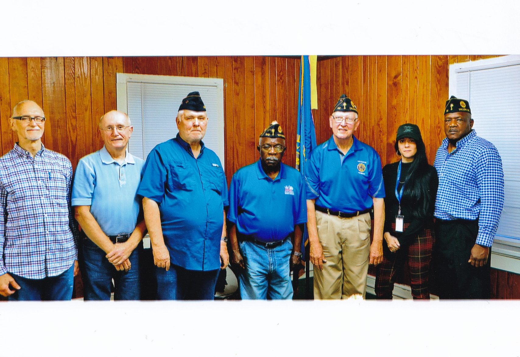 Seven people stand side by side in a room with wooden walls, some wearing military hats and blue shirts, posing for a group photo.