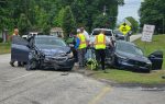 Two heavily damaged cars after a crash, emergency responders at the scene, with road signs, houses, and greenery in the background.