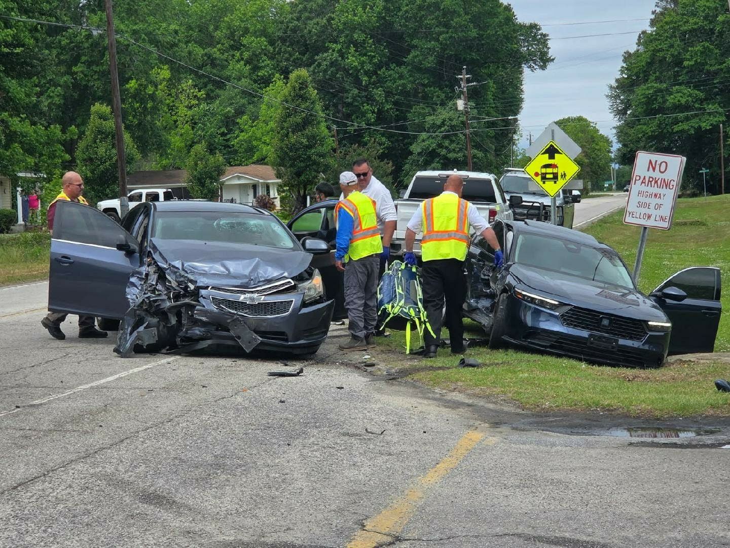 Two heavily damaged cars after a crash, emergency responders at the scene, with road signs, houses, and greenery in the background.