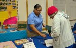 Two women interact at an educational table labeled "Magnetism" with magnets and a "Matching Game" box in a community center setting.
