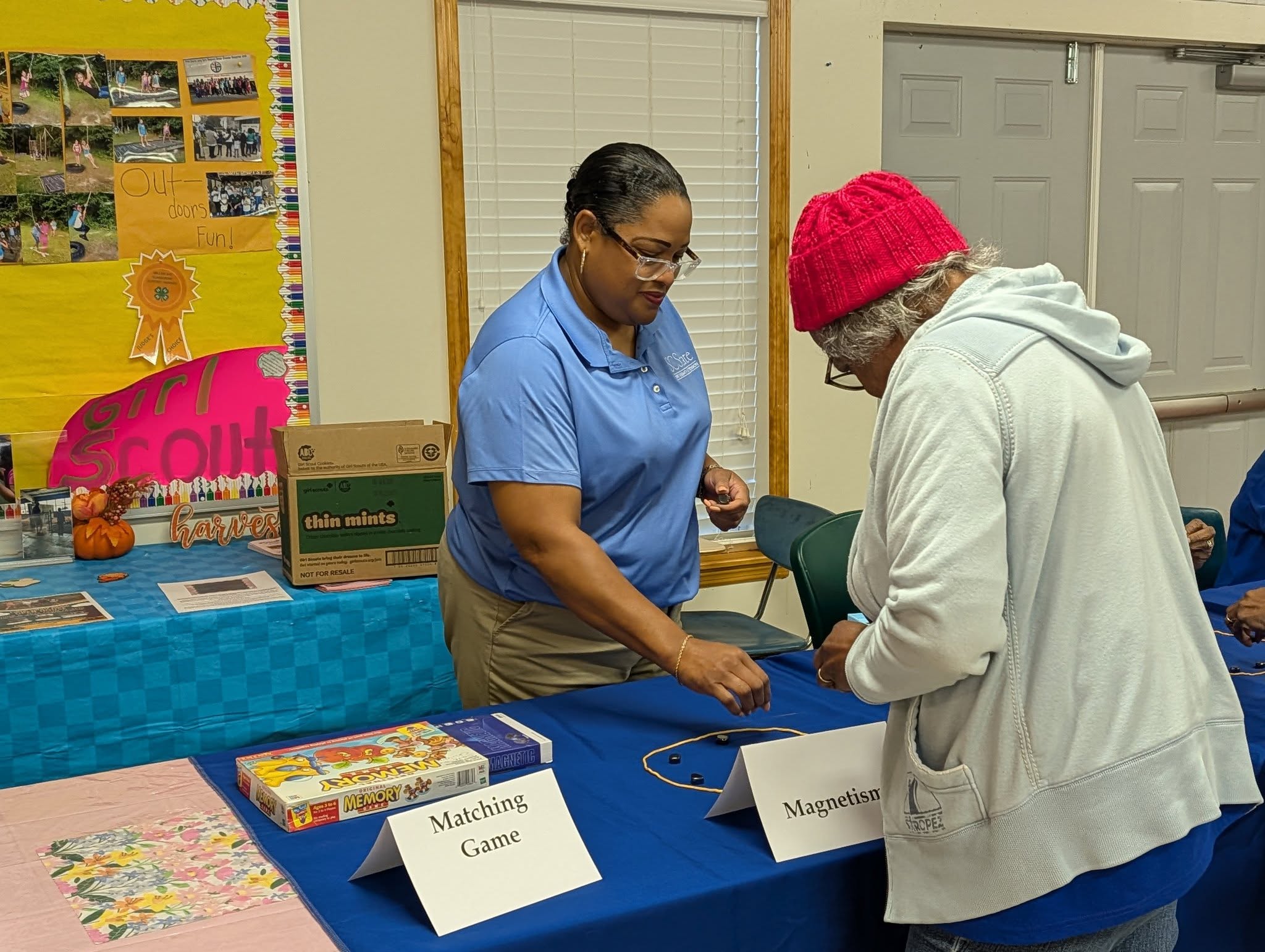 Two women interact at an educational table labeled "Magnetism" with magnets and a "Matching Game" box in a community center setting.