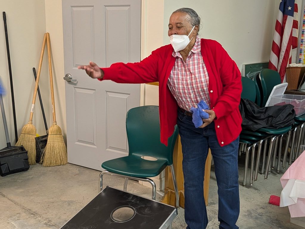 older woman wearing a mask and red sweater plays a game of cornhole indoors, surrounded by chairs, brooms, and an American flag.