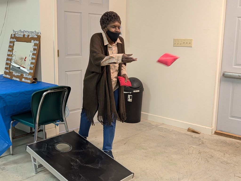 elderly woman wearing a mask plays an indoor game of cornhole, tossing a red bean bag toward the board.