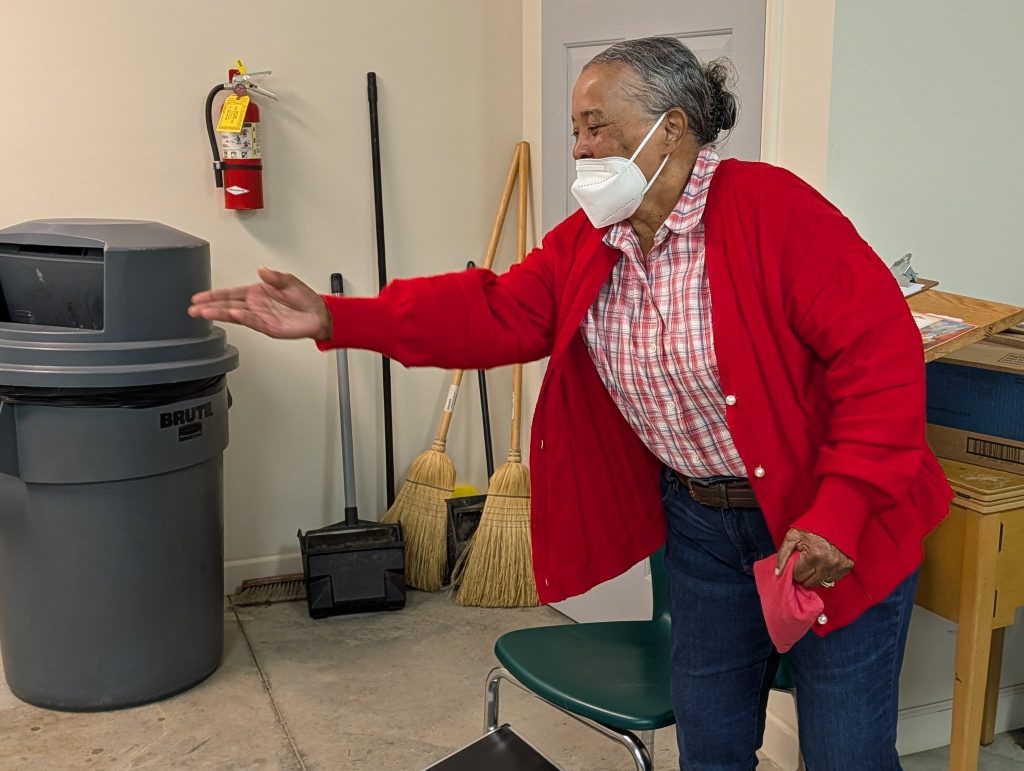 Older woman in a red sweater wearing a mask, tossing an object indoors near cleaning supplies and a trash can.