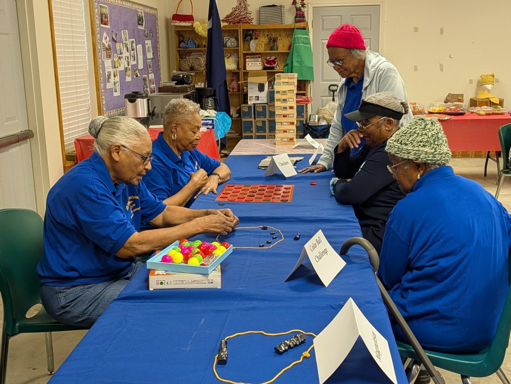 Five elderly women sit at a table playing games like checkers and a ball challenge, wearing blue shirts in a community room decorated with crafts and snacks.