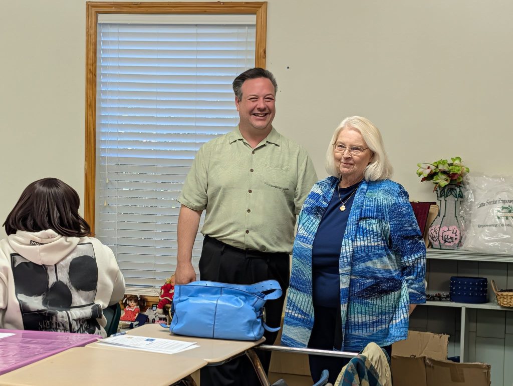 A man and an older woman stand smiling in a classroom, while another person sits at a desk in the foreground.