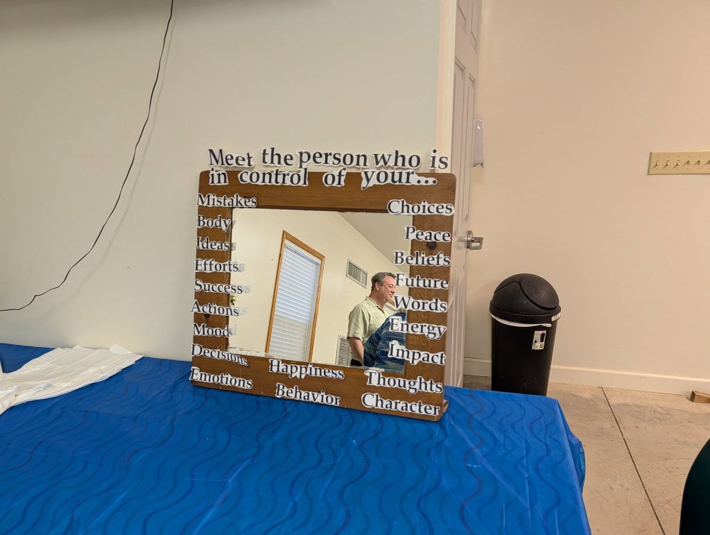 A mirror on a blue table with motivational text about self-control, reflecting a man standing in a room with white walls and a black trash can.