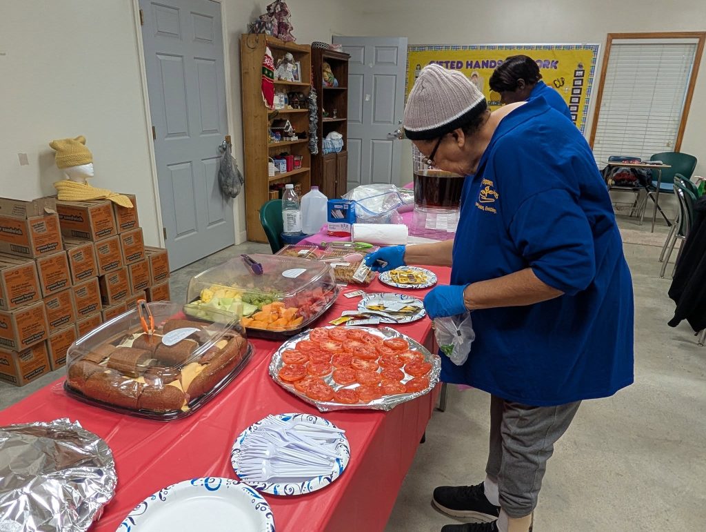 Two women in blue shirts prepare food on a table with sandwiches, fruit, and veggies in a community room. Boxes and supplies are visible in the background.