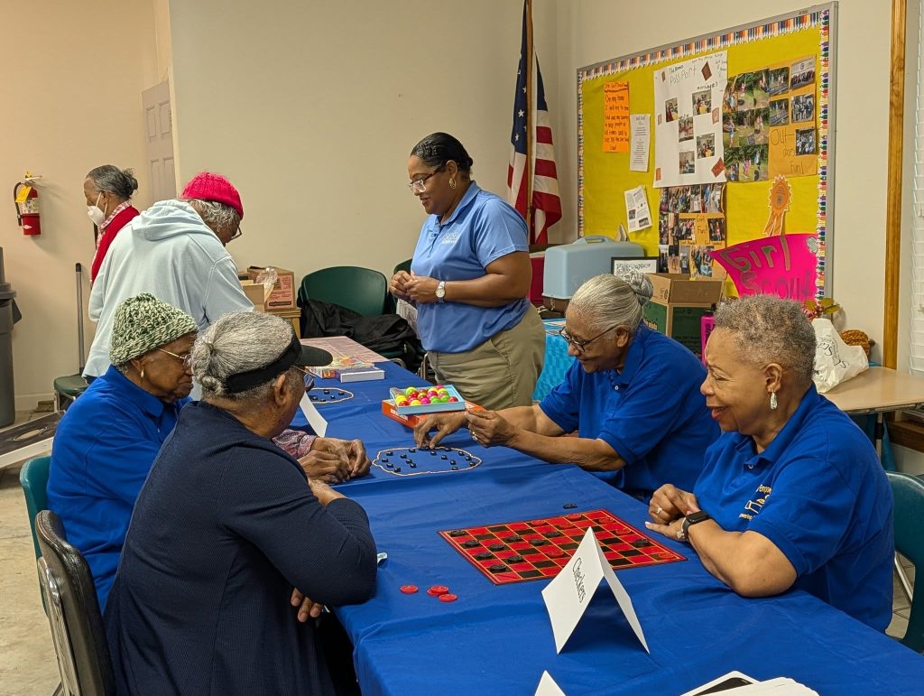 Senior women playing board games at a community center, with others standing nearby. Bulletin board and American flag in the background.