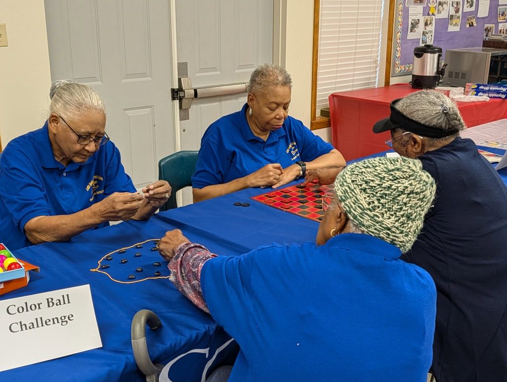 Four elderly women sitting at a table, playing board games and participating in a Color Ball Challenge, wearing blue shirts in a community room.