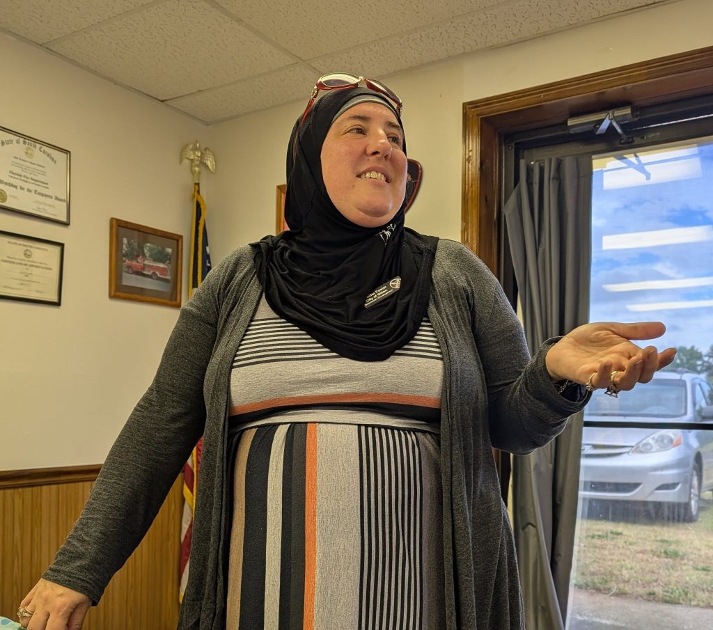 A woman in a striped dress and hijab stands inside a room, gesturing with one hand, with certificates and an American flag in the background.