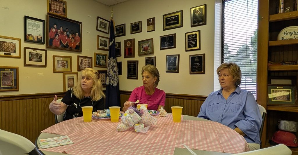 Three women sit at a round table with food and drinks, in a room decorated with award plaques, framed photos, and a flag.