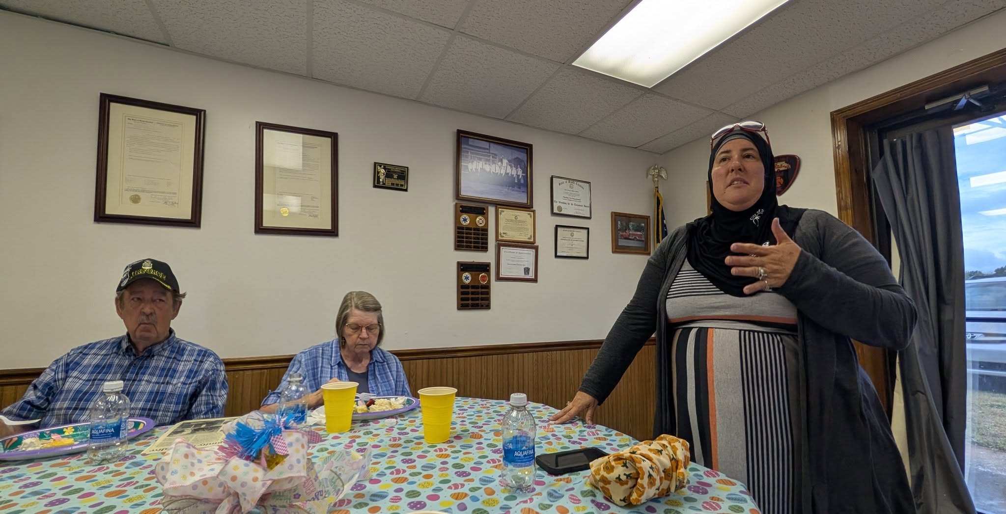 Three people at a table with food and drinks, one woman standing and speaking, wall behind decorated with framed certificates and plaques.